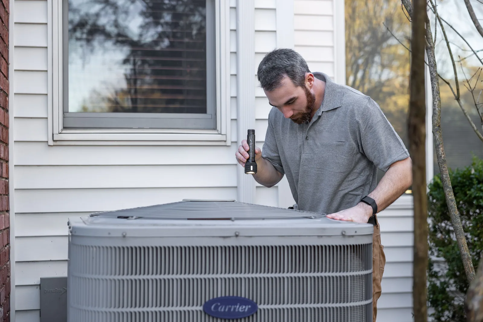 HVAC technician inspecting an outdoor air conditioning condenser beside a home during AC maintenance